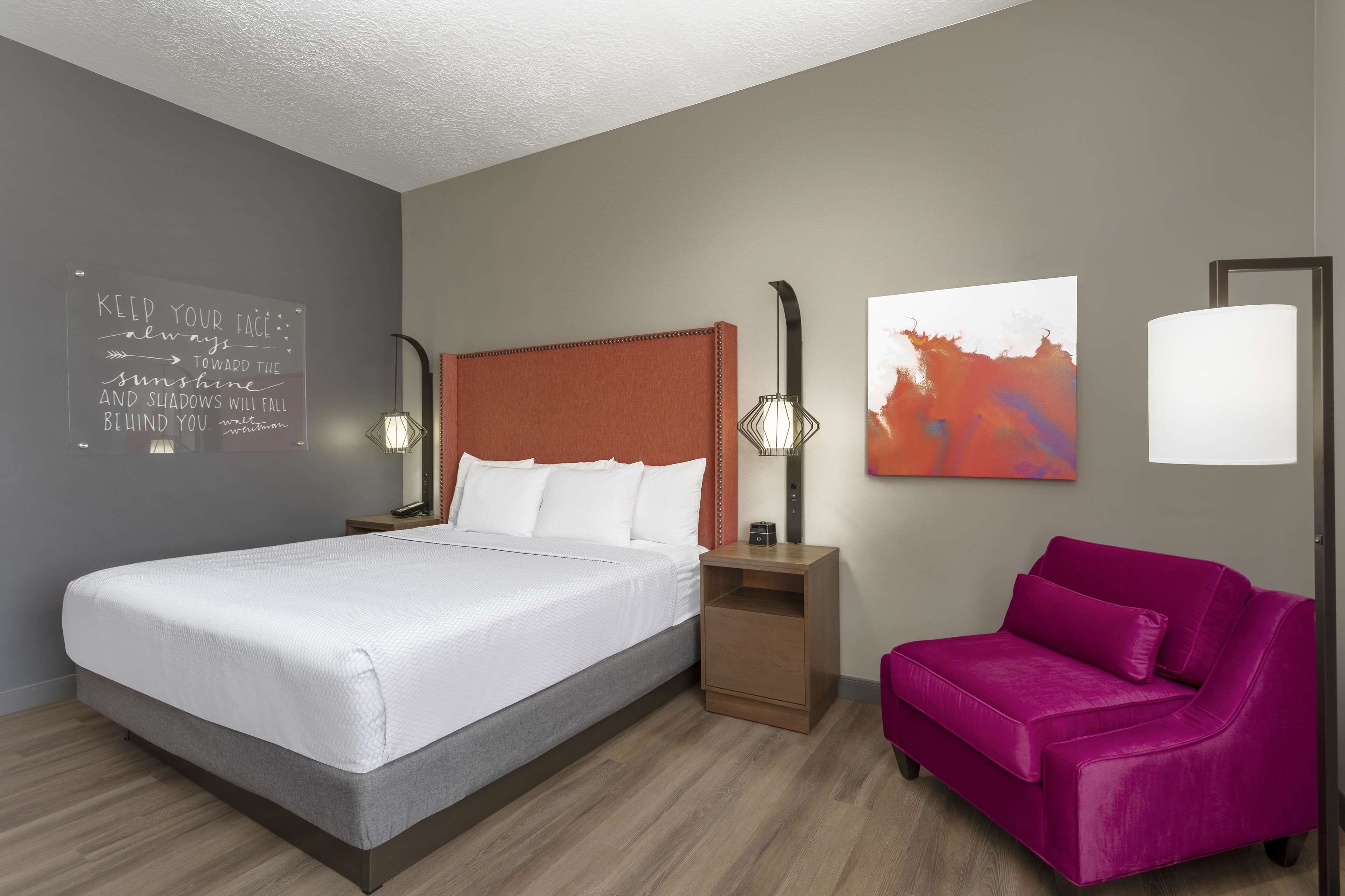 A hotel room with a large bed, a nightstand, a pink chair, a wall art piece, and a motivational quote on the wall.