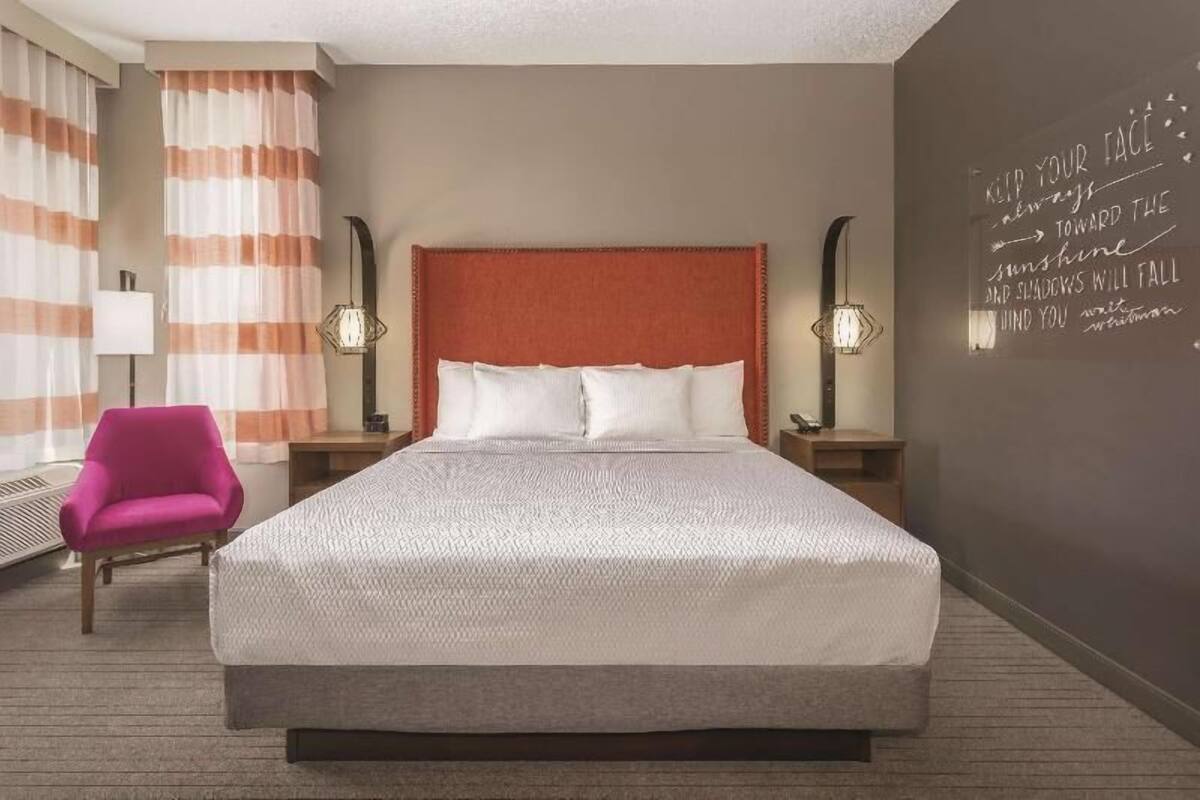 A hotel room with a large bed, a pink chair, two bedside tables with lamps, and a wall with a motivational quote.