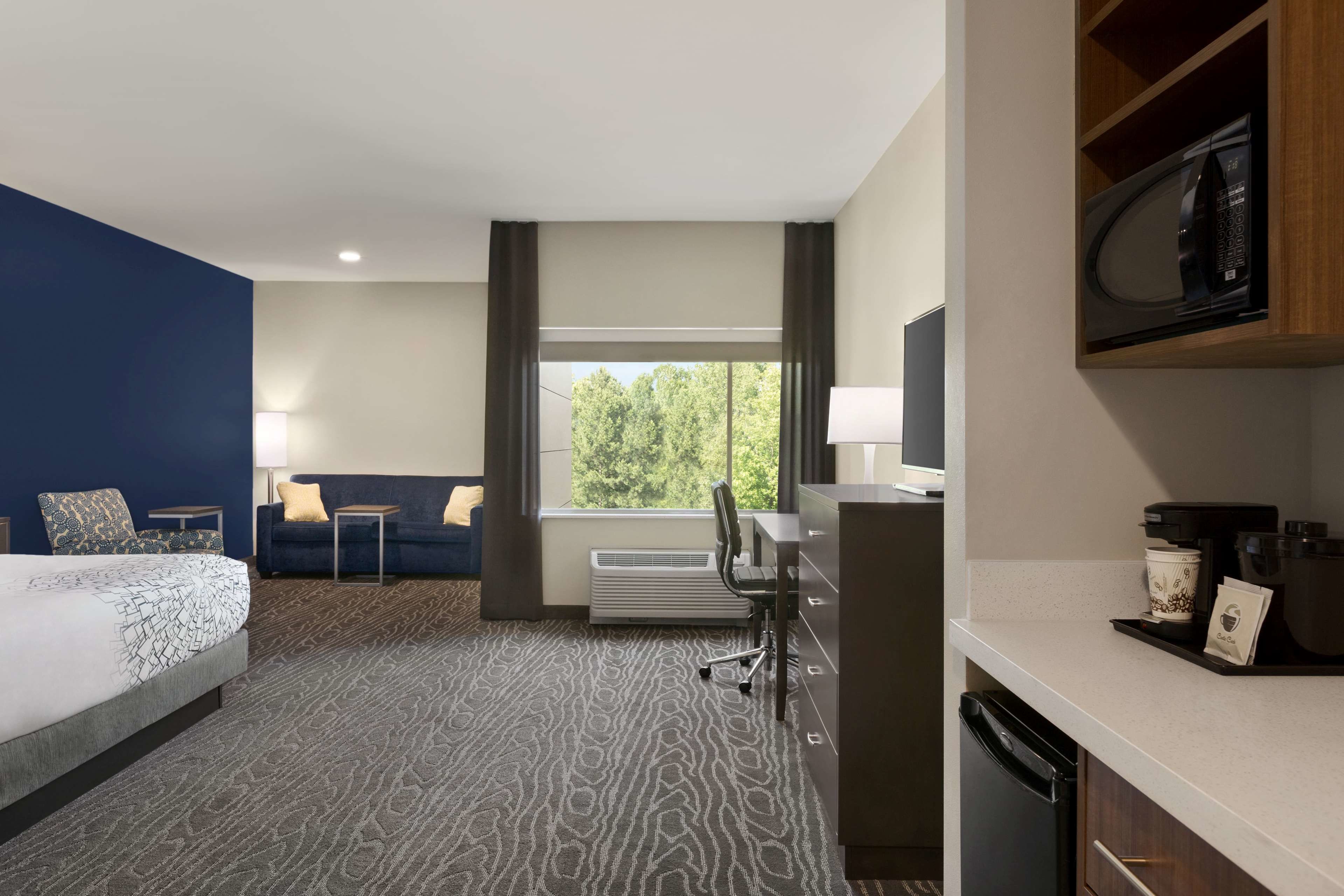 A hotel room with a bed, a desk, a chair, a microwave, a coffee maker, and a view of trees through the window.