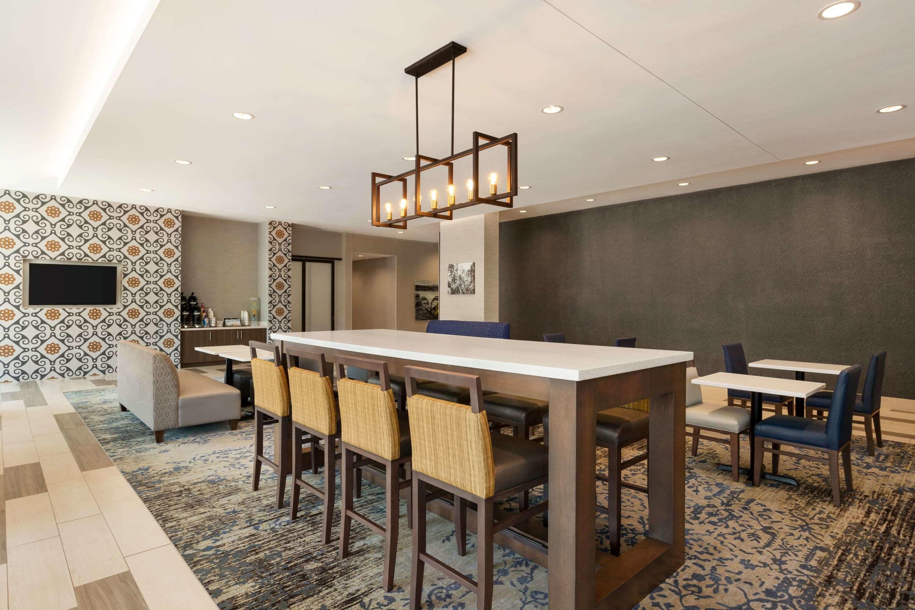 A modern hotel lobby with a long reception desk, multiple seating areas, patterned walls, and a ceiling with recessed lighting.