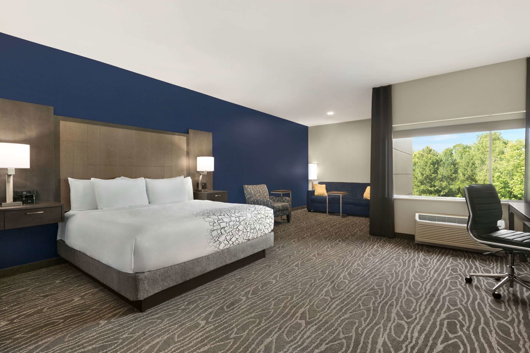 Habitación de hotel con una cama grande, un escritorio, una silla y vistas a árboles por la ventana.