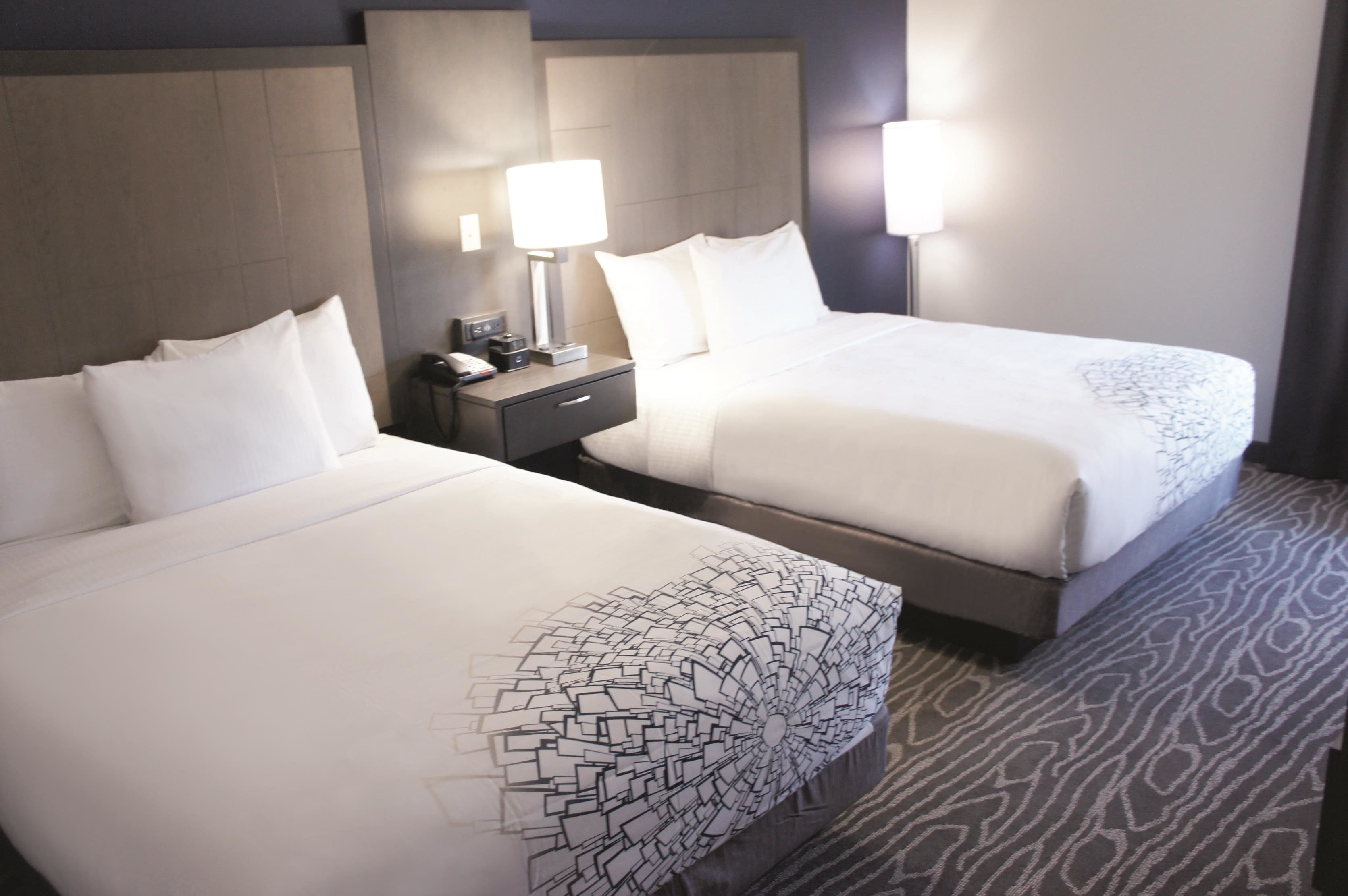 Two beds with white bedding and a patterned pillow, a nightstand with a lamp, and a patterned carpet.