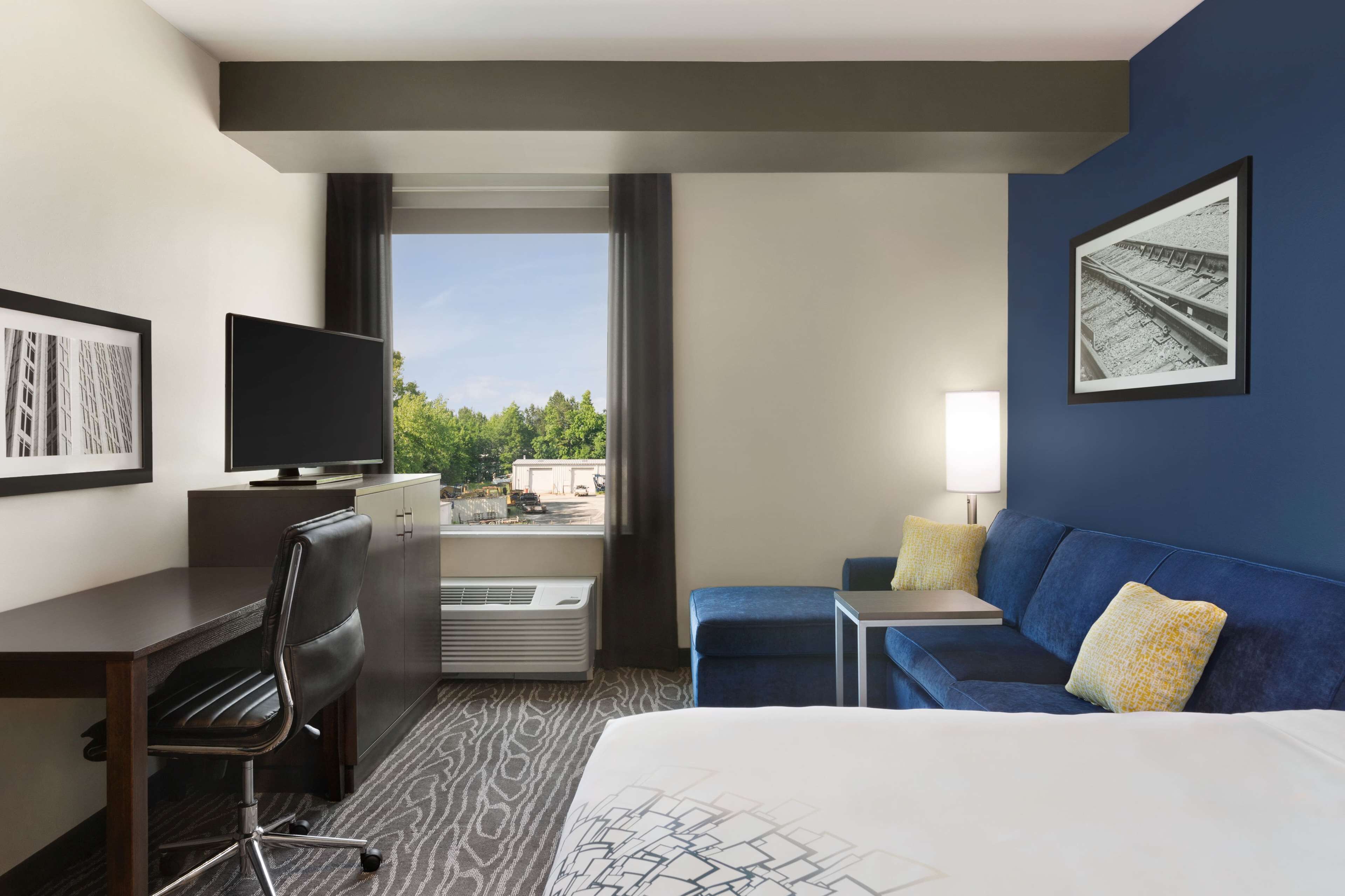 Habitación de hotel con cama, escritorio con silla, televisor, sofá y ventana con vistas a zonas verdes.