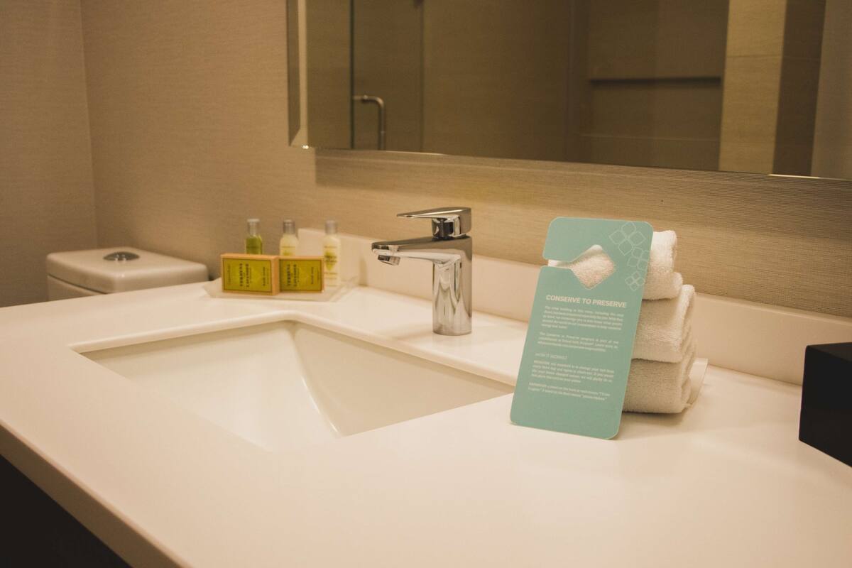A hotel bathroom with a white sink, a mirror, and a towel rack. There are toiletries on the sink counter.