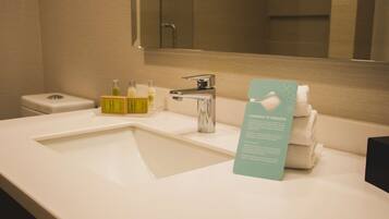 Un baño de hotel con un lavabo blanco, un espejo y un perchero. Hay artículos de aseo en la encimera del lavabo.