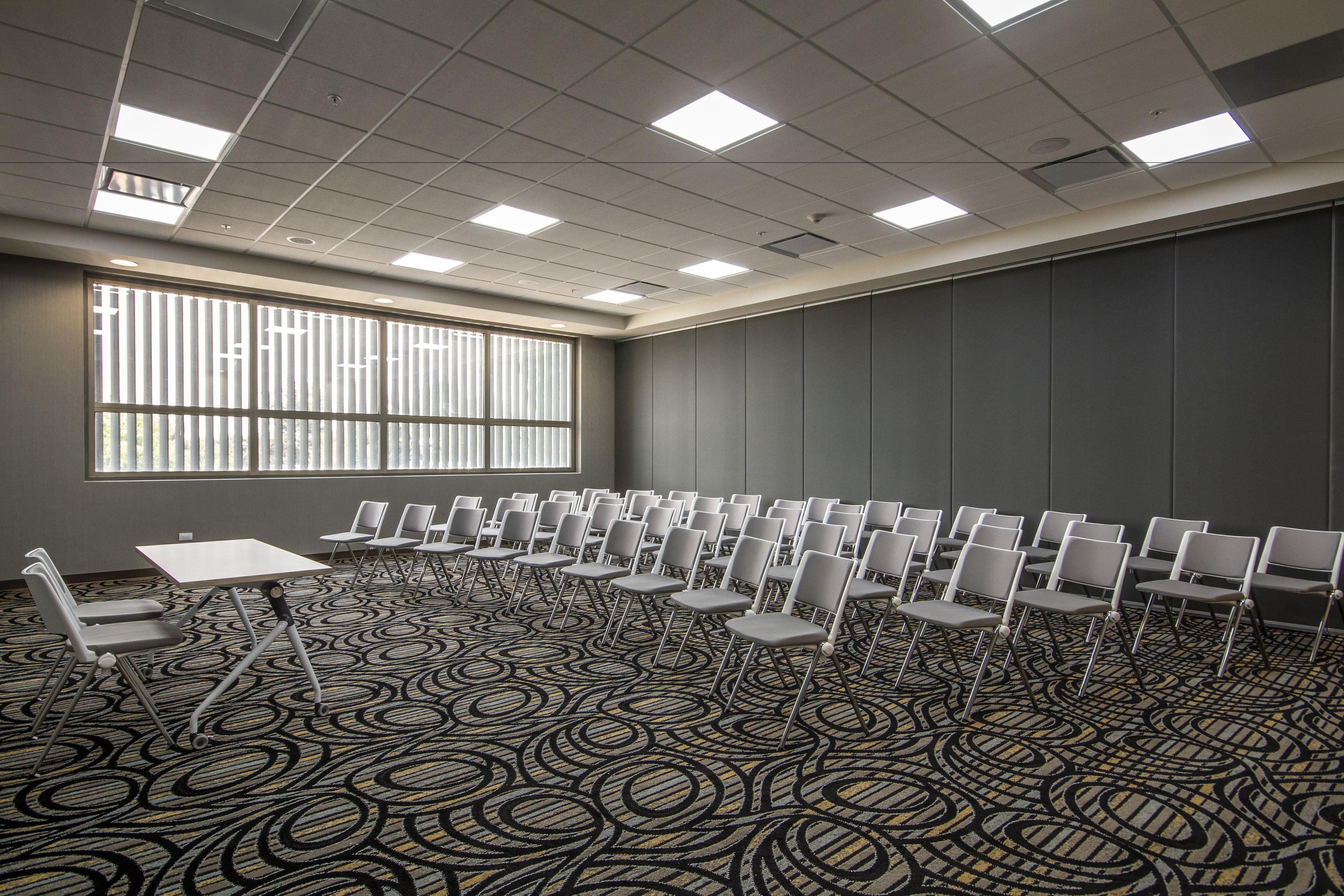 Un salón de conferencias con alfombra estampada, hileras de sillas, una mesa y un ventanal con persianas.