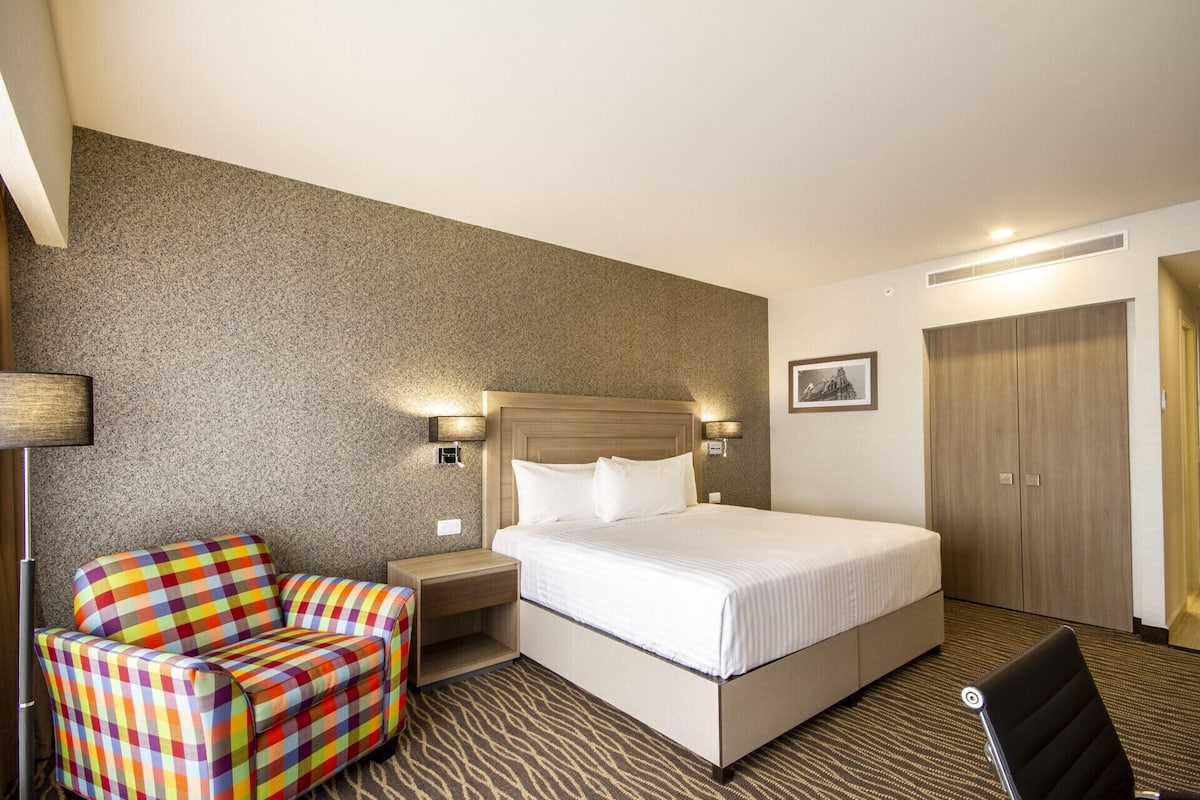 A hotel room with a bed, a chair, a nightstand, a lamp, and a wardrobe.