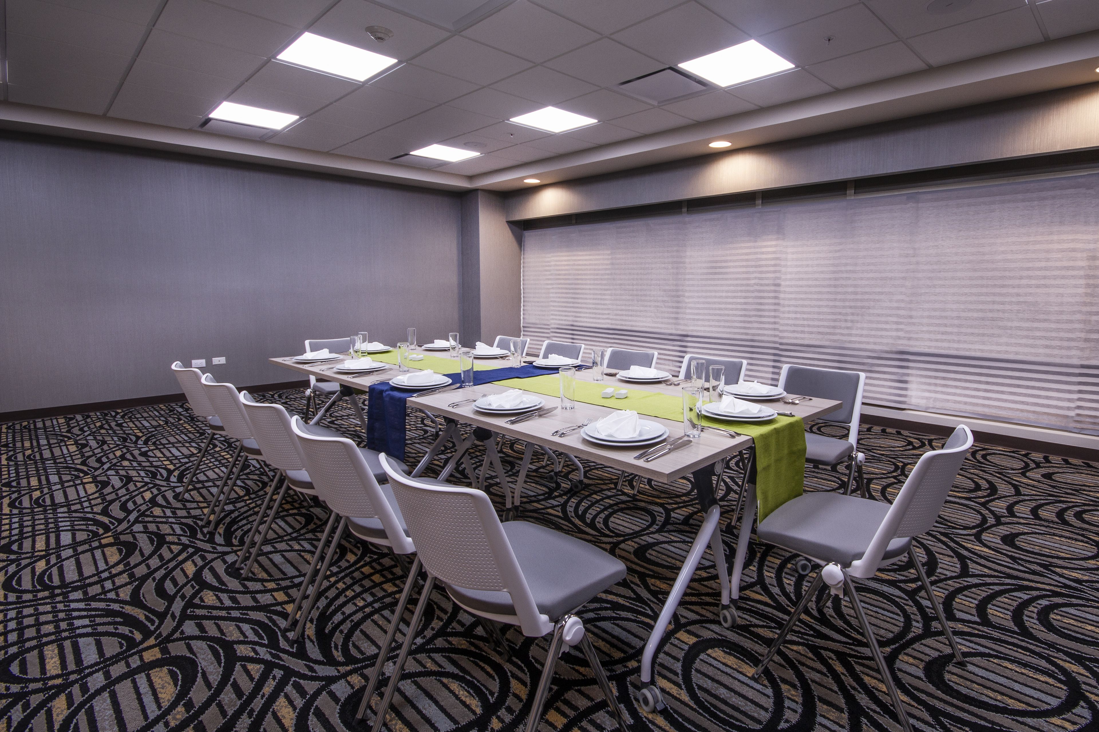 A conference room with a long table set for a meeting, surrounded by chairs, and a large window with blinds.