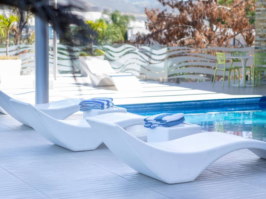 Área de piscina com espreguiçadeiras brancas e uma pequena mesa com toalhas.