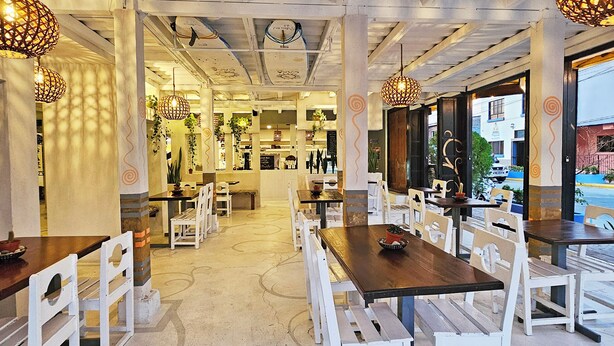 Een eetruimte binnenshuis met witte stoelen en tafels, decoratieve hanglampen en uitzicht op de straat.