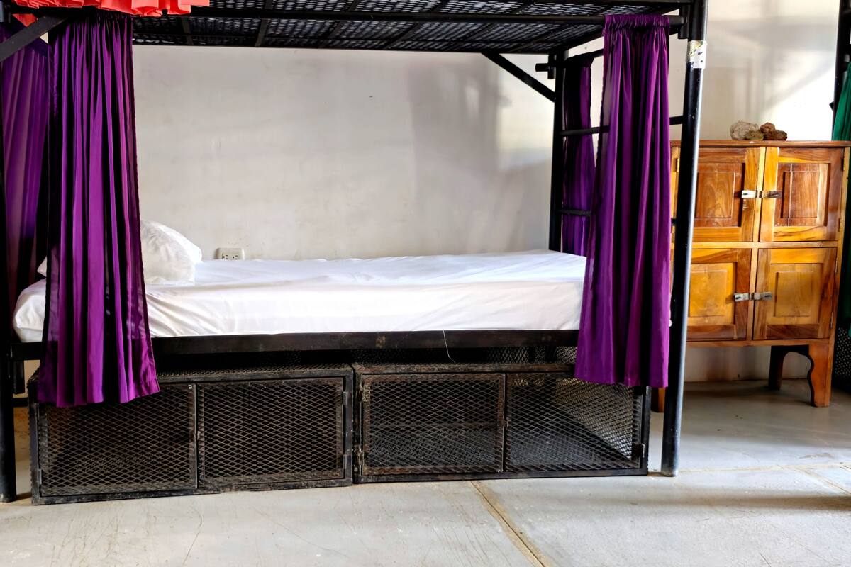 Un lit simple avec un baldaquin, une armoire en bois et un ventilateur sur pied.