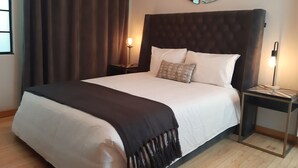Una cama bien tendida con una colcha negra, una lámpara de noche y un espejo en la pared.