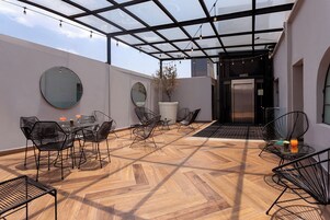 Une terrasse extérieure dotée d’un sol en bois disposé en motif de chevron, de chaises en métal noir et d’une structure de toit en verre.