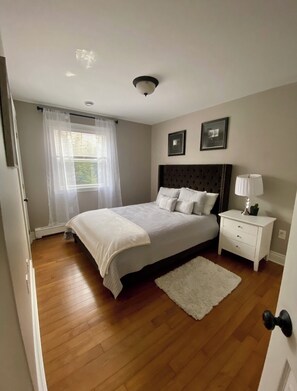 Un dormitorio con una cama grande, una mesita de noche con lámpara, una alfombra blanca y una ventana con cortinas de encaje.