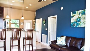 A modern living room with a dark brown leather sofa, a round coffee table, and a blue rug. There are three wooden chairs and a kitchen area with pendant lights. - "The Wedgewood"/A Little Bit Country, Close To Town, New, Clean And Fresh. (Kyle)