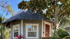 A small, single-story house with a grey exterior and a red door, surrounded by lush tropical plants.