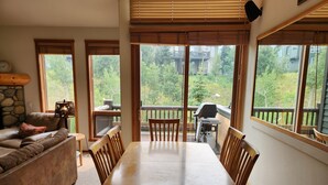 A dining area with a table and chairs, large windows with blinds, a stone wall, and a view of a balcony with a grill. - Snowcreek Resort Vacation Rental 954 (Mammoth Lakes)