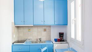 A compact kitchen with blue cabinets, a wooden countertop, a coffee maker, and a small dining table with a chair. - Myconian Brothers    (mykonos)