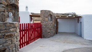 A stone wall with a red gate, a white building with a red door, and a view of a hill with white houses. - Myconian Brothers    (mykonos)