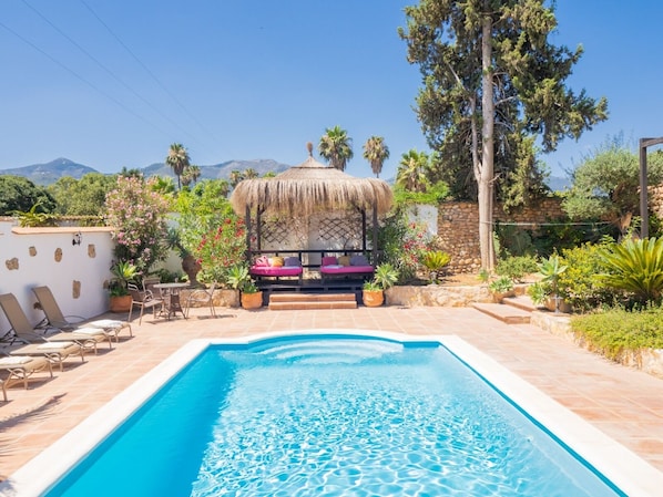 A swimming pool with lounge chairs and a thatched-roof seating area surrounded by plants and trees.