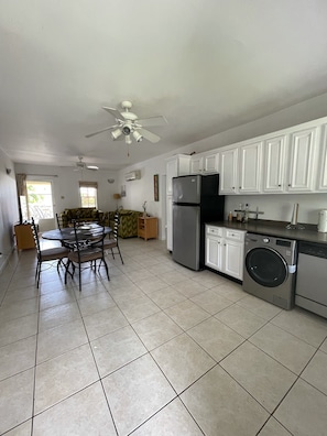 A spacious kitchen with white cabinets, a refrigerator, a washing machine, and a dining area with a round table and chairs.