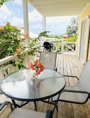 A covered outdoor patio with a round glass table, two chairs, a grill, and a white fence.