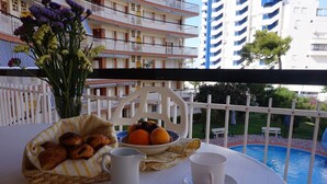 Un balcón con una mesa preparada para el desayuno, un jarrón de flores y vistas a una piscina y edificios.