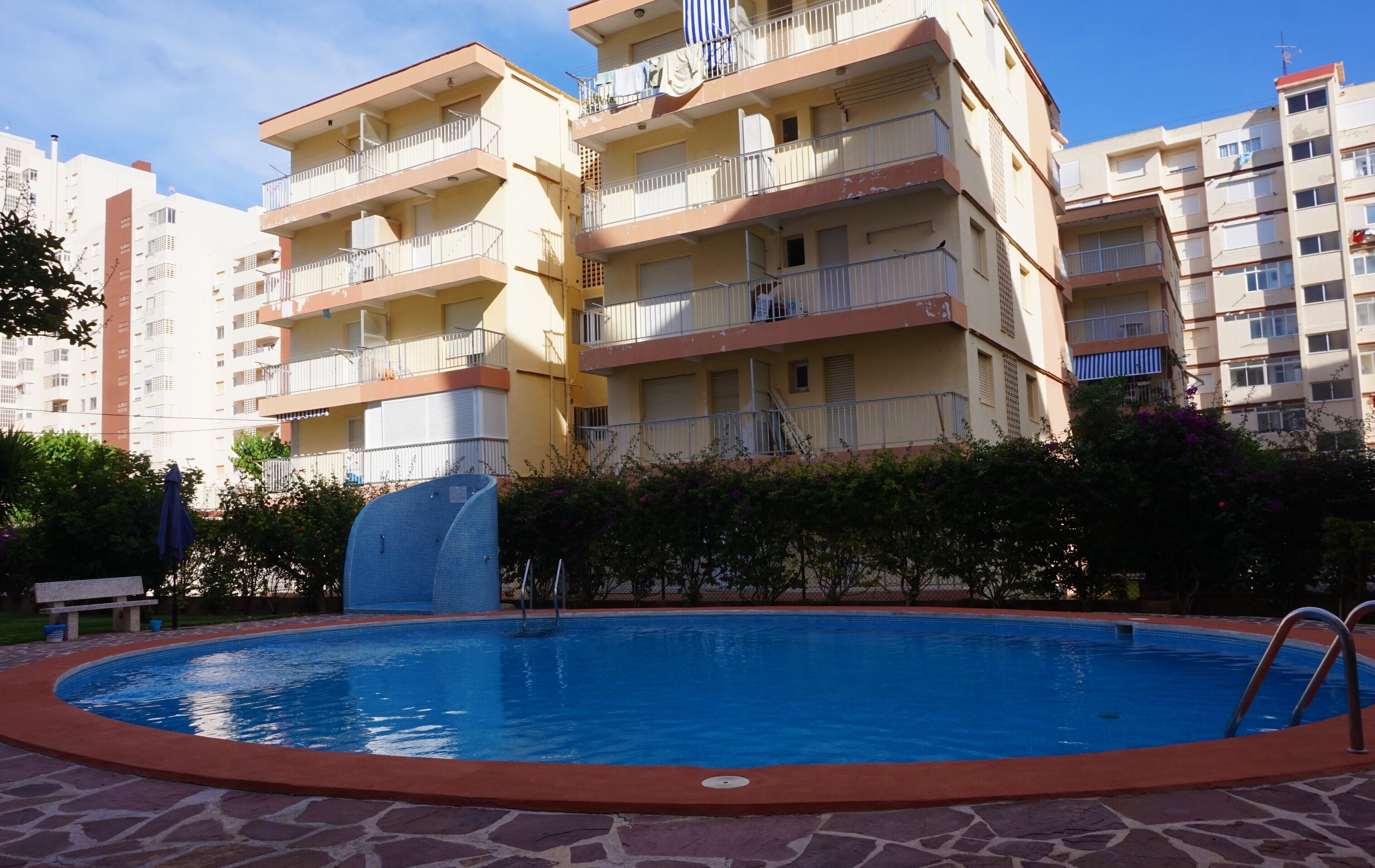 A swimming pool surrounded by a stone deck, with a building featuring multiple balconies in the background.