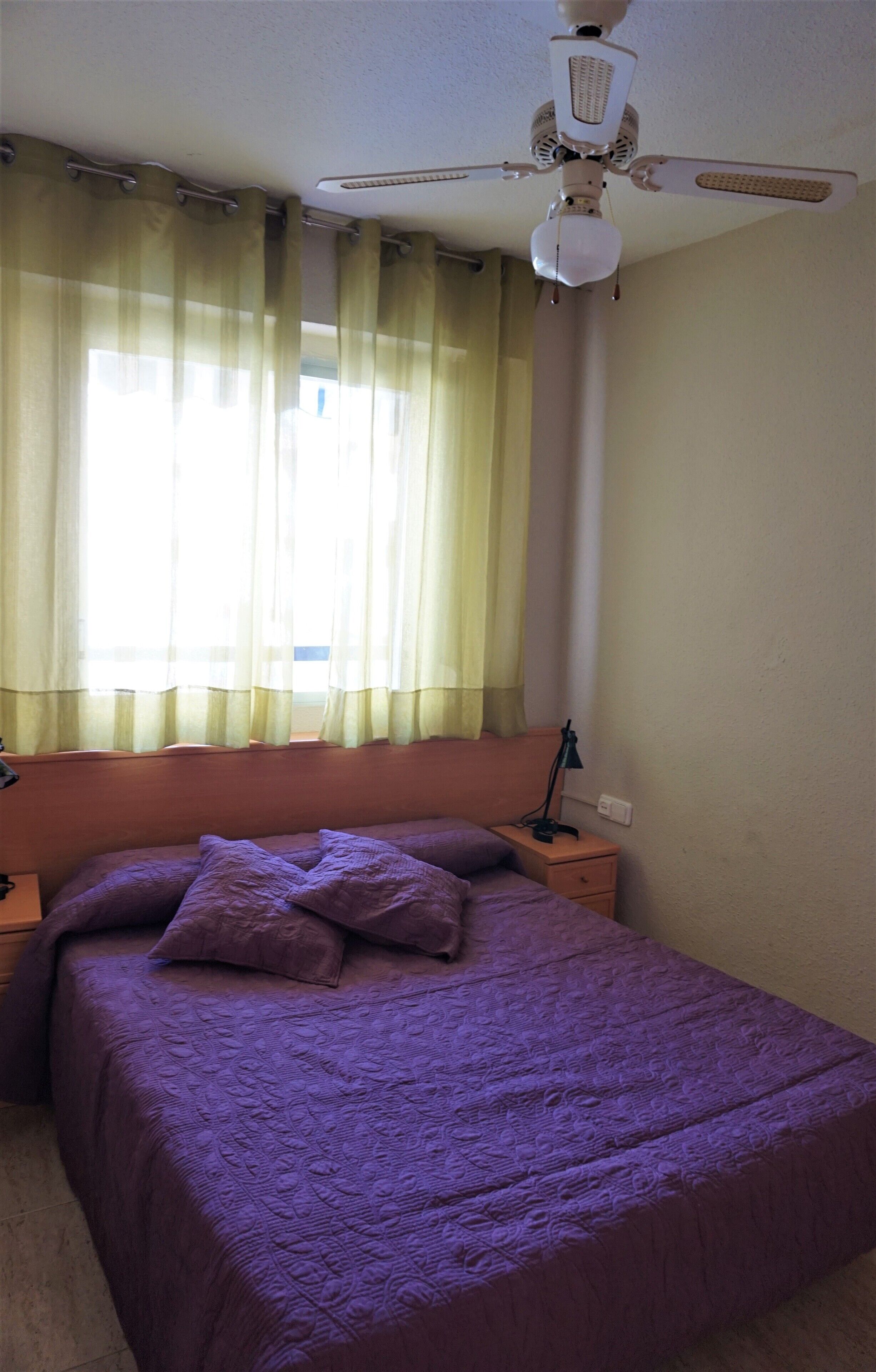 A bedroom with a bed, a nightstand, a ceiling fan, and a window with curtains.