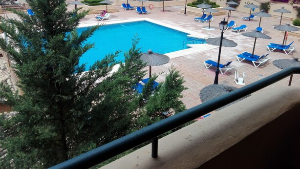 Zona de piscina con sillas y sombrillas azules, rodeada por un edificio con balcones y vistas despejadas a la piscina.