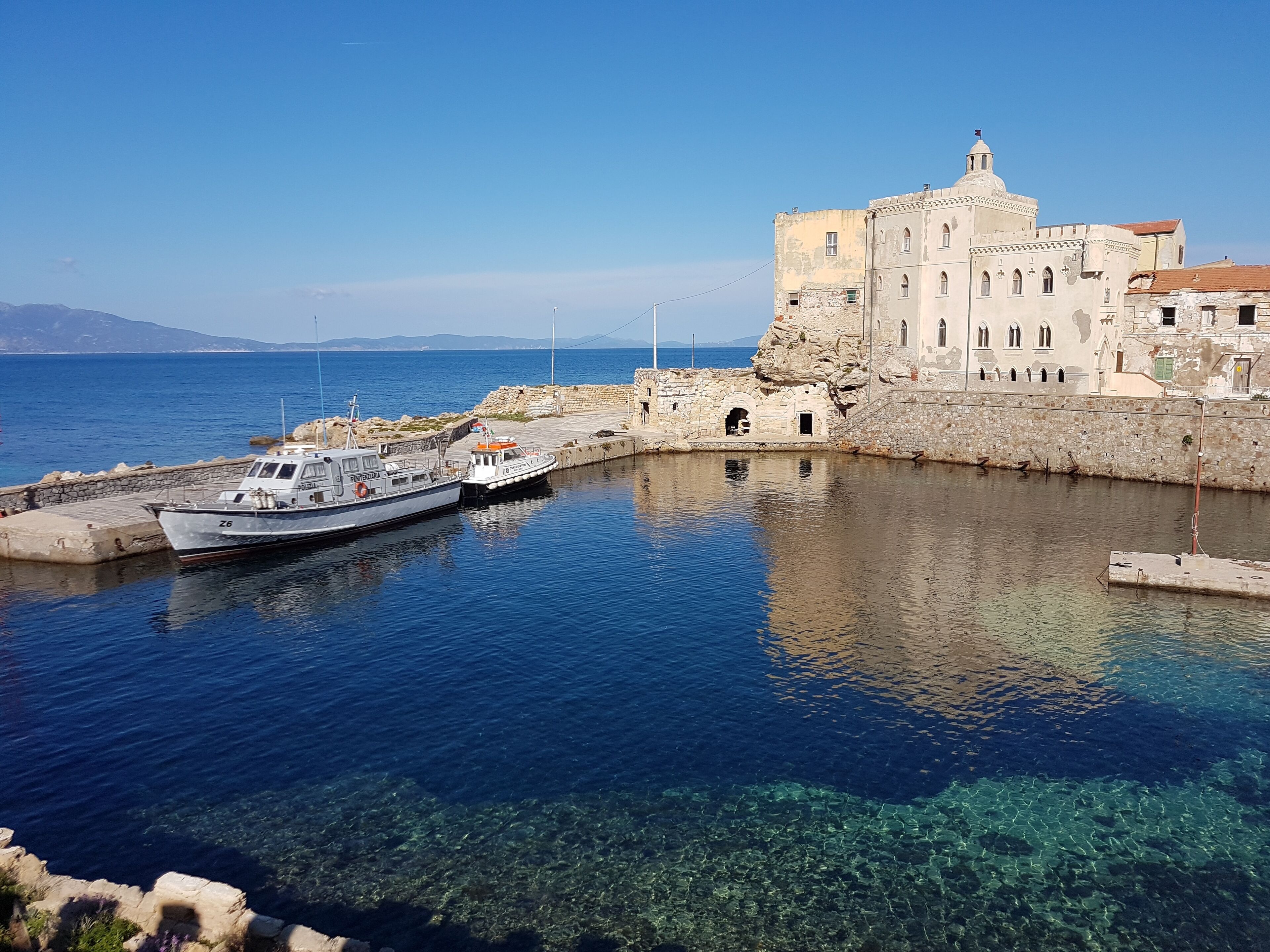 Un puerto con barcos amarrados, un edificio de piedra con ventanas de arco y agua clara que refleja el cielo.