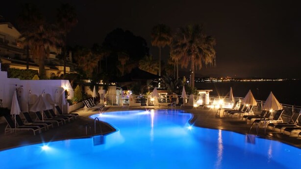 Une piscine nocturne, dotée de chaises longues et de parasols, entourée de palmiers et offrant une vue sur le paysage urbain.