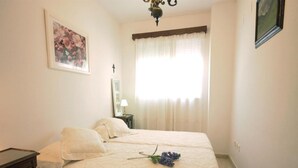 Quarto com cama, mesa de cabeceira, abajur, quadro emoldurado e janela com cortinas.