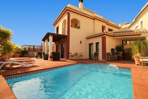 A pool area with a clear blue pool, a covered patio, and a building with a tiled roof and arched windows.