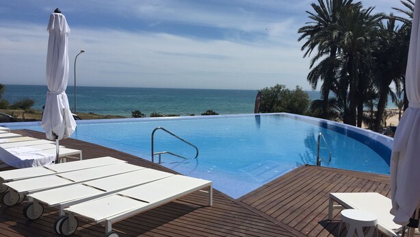 Une piscine à débordement avec une terrasse en bois, des chaises longues et des parasols.