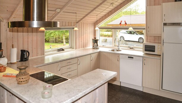 A modern kitchen with wooden cabinets, a stainless steel range hood, a built-in dishwasher, and a window with a view of a white car and greenery outside. - Nice home in Oksbøl with sauna (Oksbøl)
