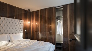 Une chambre à coucher avec une tête de lit capitonnée, des murs recouverts de panneaux de bois, un dressing avec des vêtements suspendus et un lit avec du linge de lit blanc.