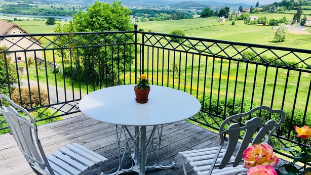 Un balcon avec une table ronde et deux chaises, donnant sur un paysage verdoyant où l’on aperçoit une église et des maisons.