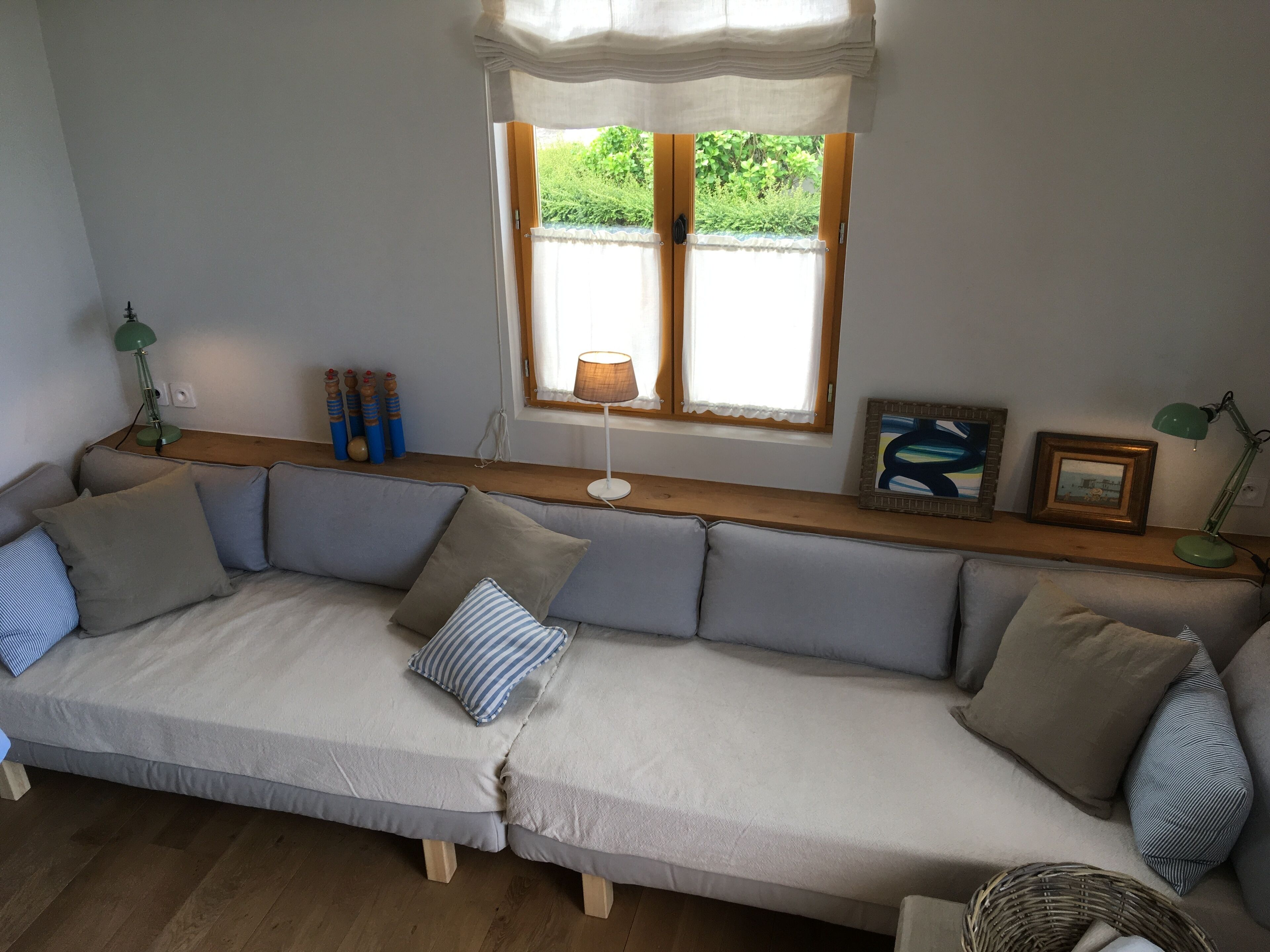 A cozy living room with a large L-shaped sofa, a small table with decorative items, a window with curtains, and a lamp on the table.