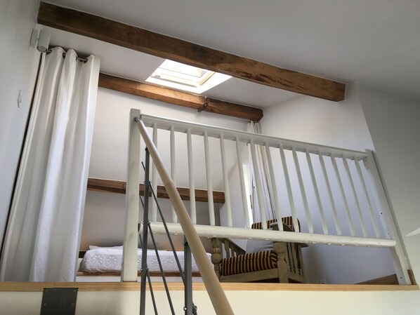 A loft bedroom with a skylight, a wooden beam ceiling, a white railing, a bed with a white and patterned bedspread, and a striped chair.
