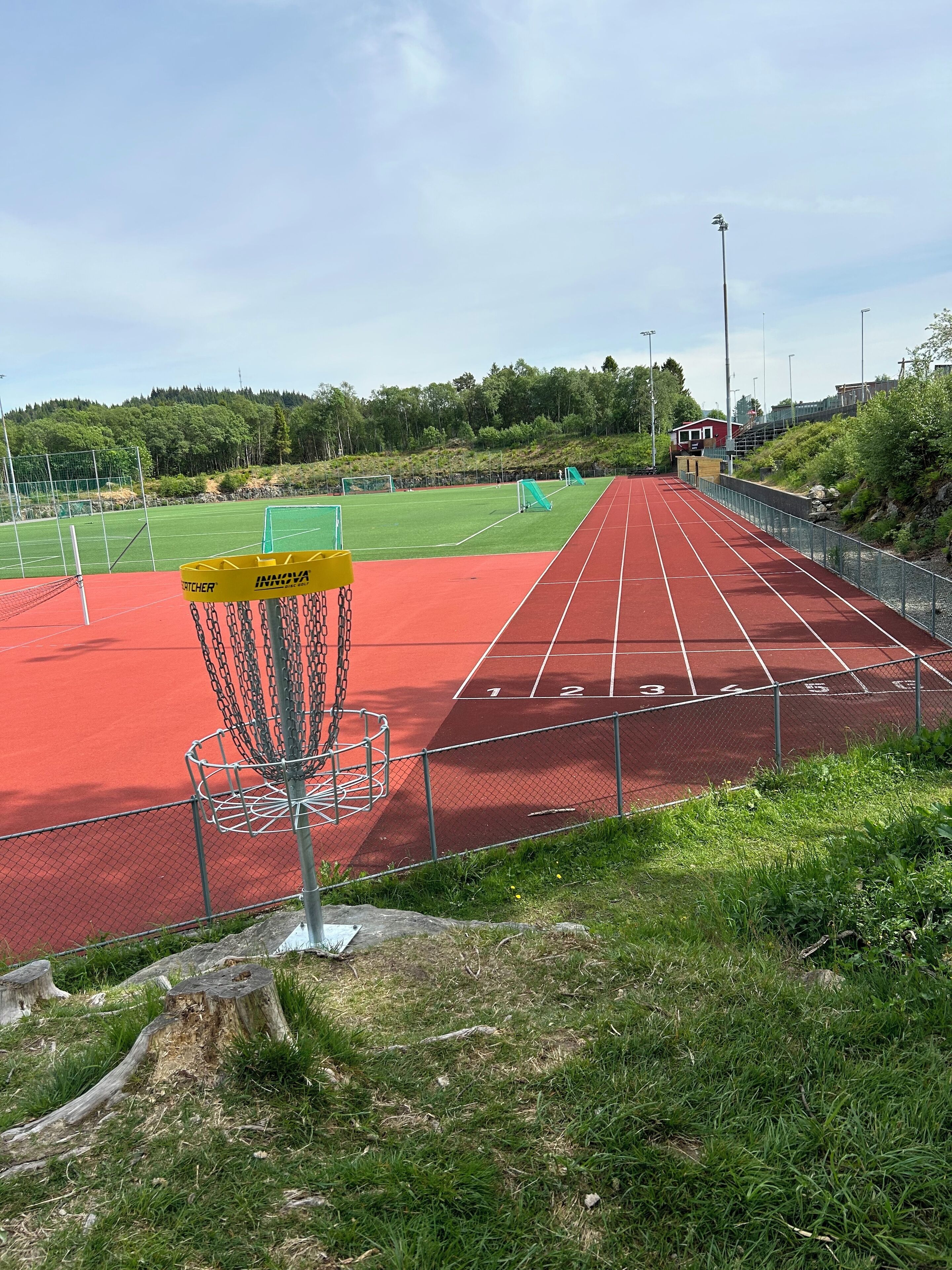 A red running track with a chain-link fence, a yellow disc golf basket, and a green field in the background.