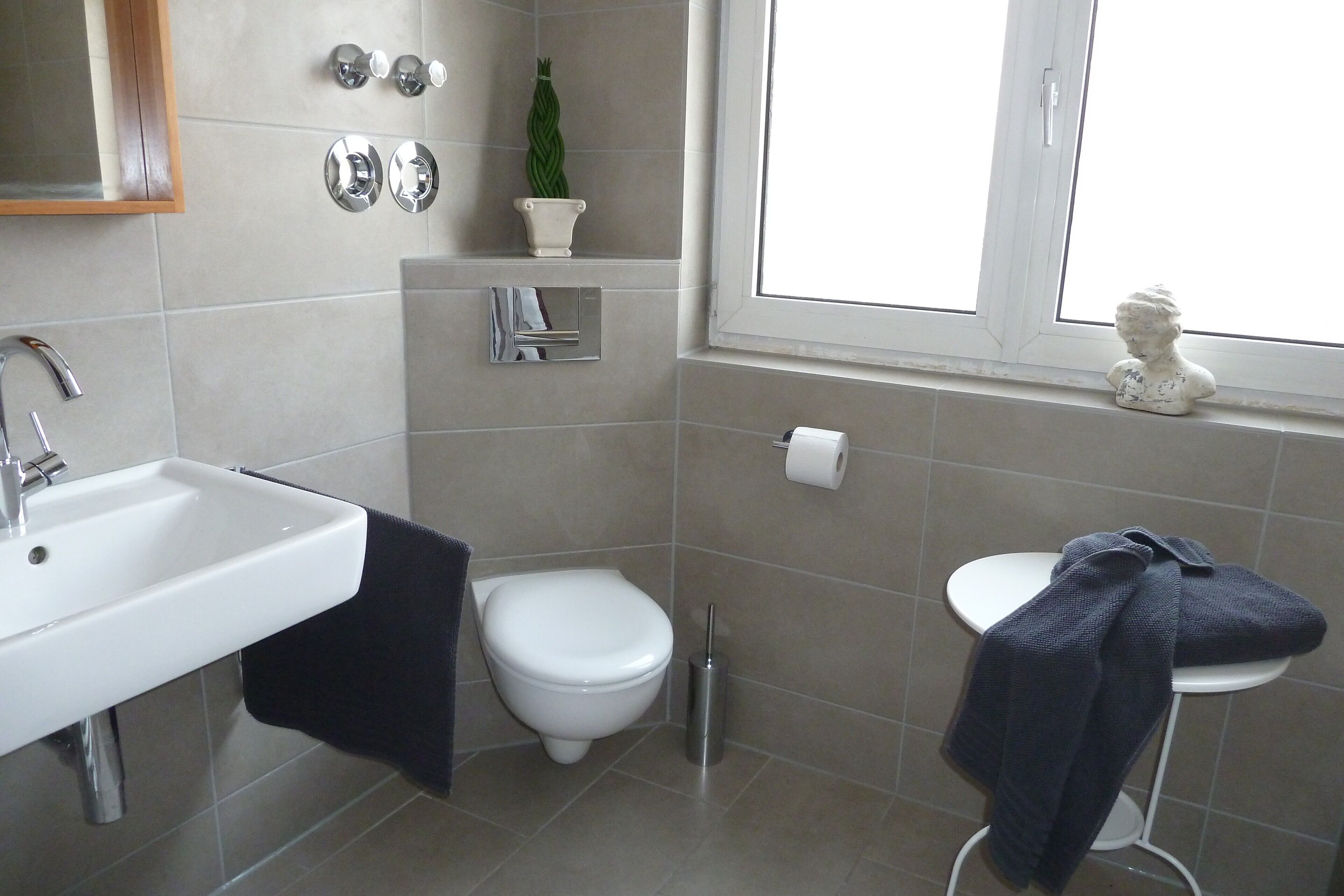Une salle de bain avec un lavabo, des toilettes et un bidet, tous de couleur blanche. Il y a une fenêtre donnant sur l’extérieur, une petite étagère avec une plante et une statue posée sur le rebord de la fenêtre.