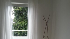 Una habitación con piso de madera, un aparador blanco, un tapete negro y una ventana con cortinas blancas.