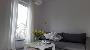 Una sala de estar con un sofá gris, una mesa de centro blanca, un jarrón con flores amarillas y una ventana con cortinas blancas.