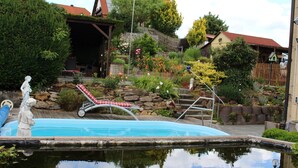Une piscine ornée d’une statue, un fauteuil de jardin à carreaux rouges et blancs et un mur de pierre agrémenté de plantes.
