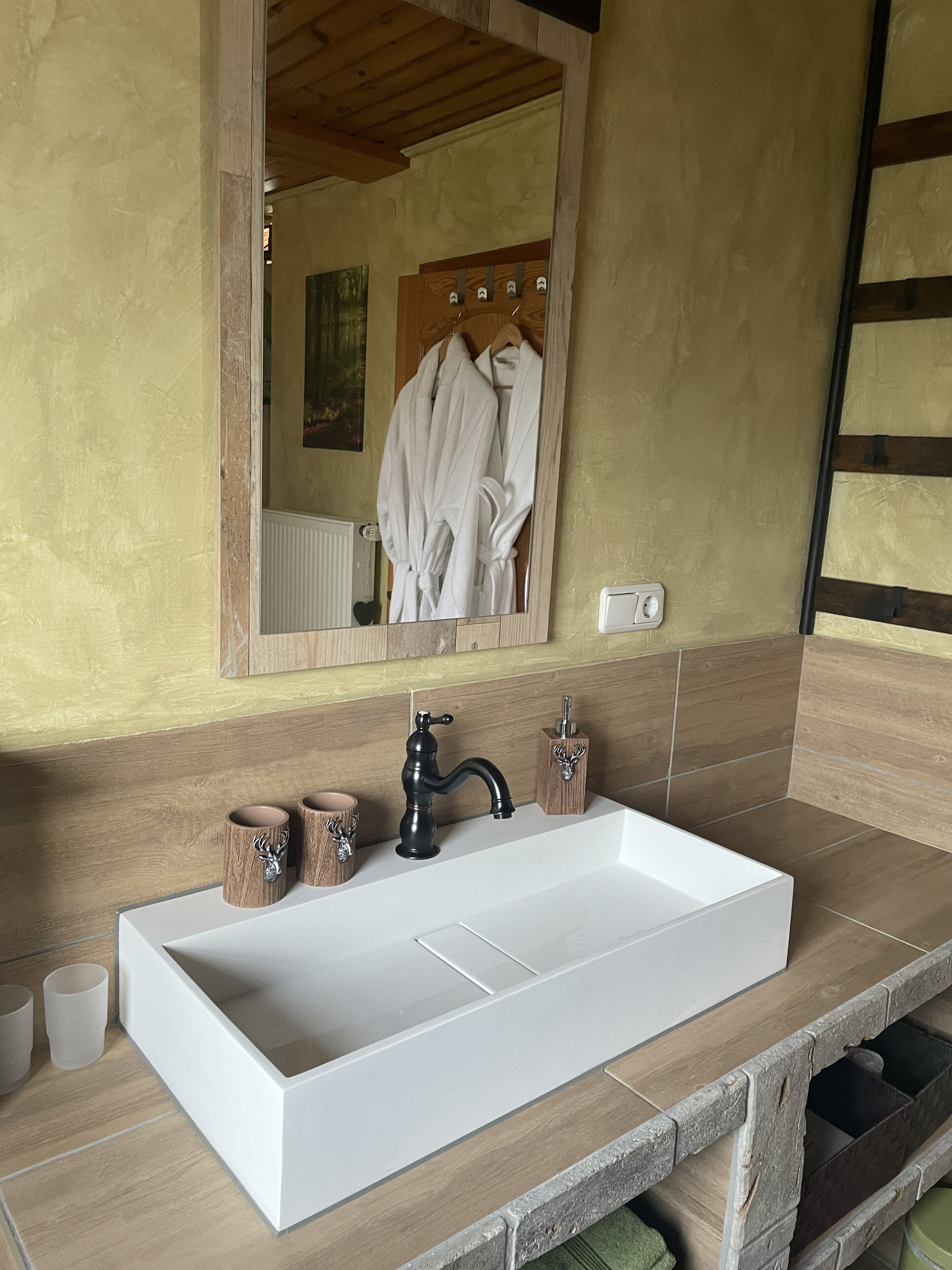 Een badkamer met een groot wit wastafel, houten wastafelmeubel en een spiegel die een badjas en een radiator weerspiegelt.