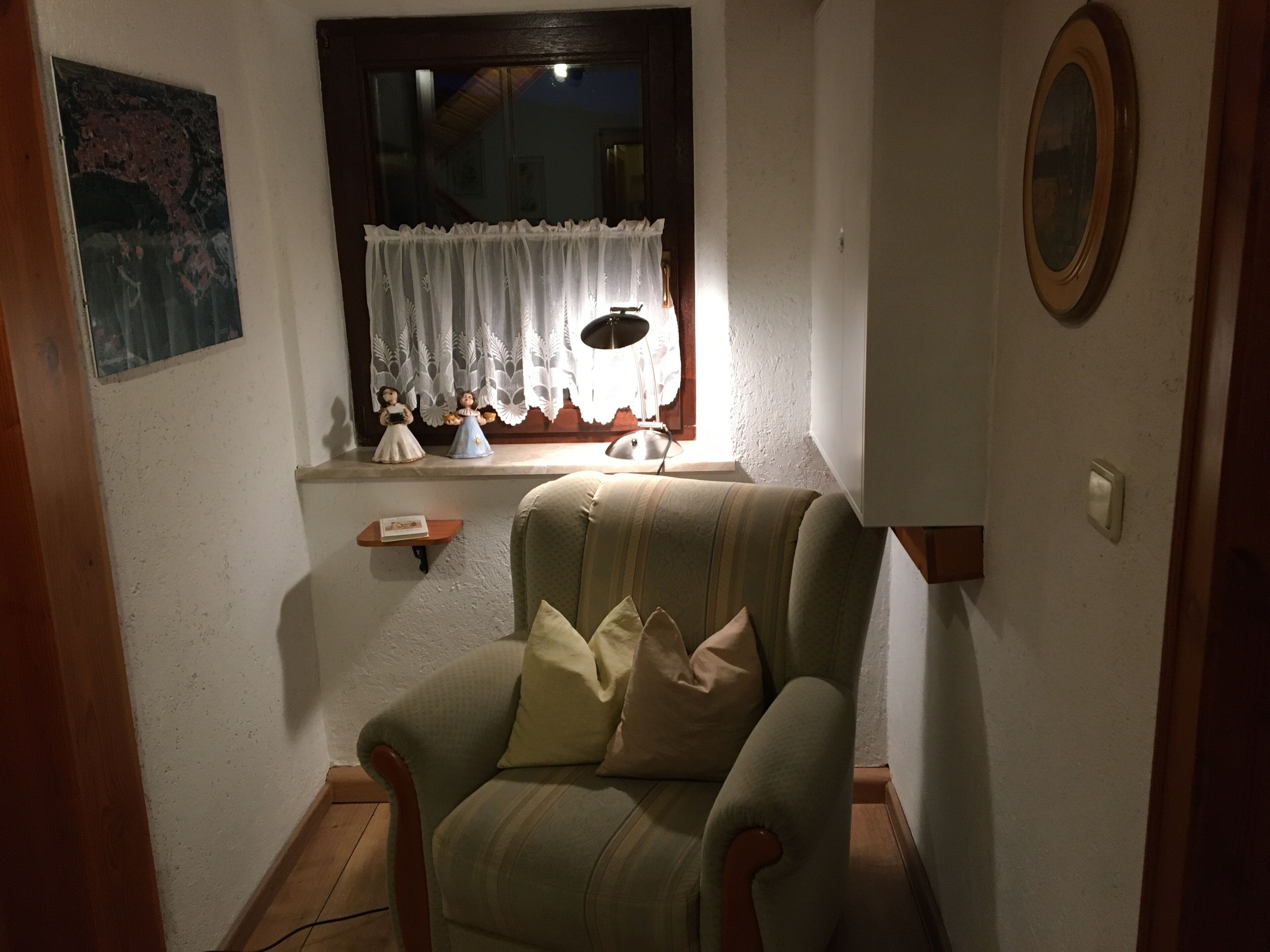 Ein gemütliches Zimmer mit einem grünen Sessel, einer kleinen Ablage mit Figuren und einem Fenster mit weißem Vorhang.