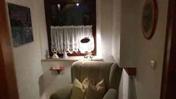 Ein gemütliches Zimmer mit einem grünen Sessel, einer kleinen Ablage mit Figuren und einem Fenster mit weißem Vorhang.