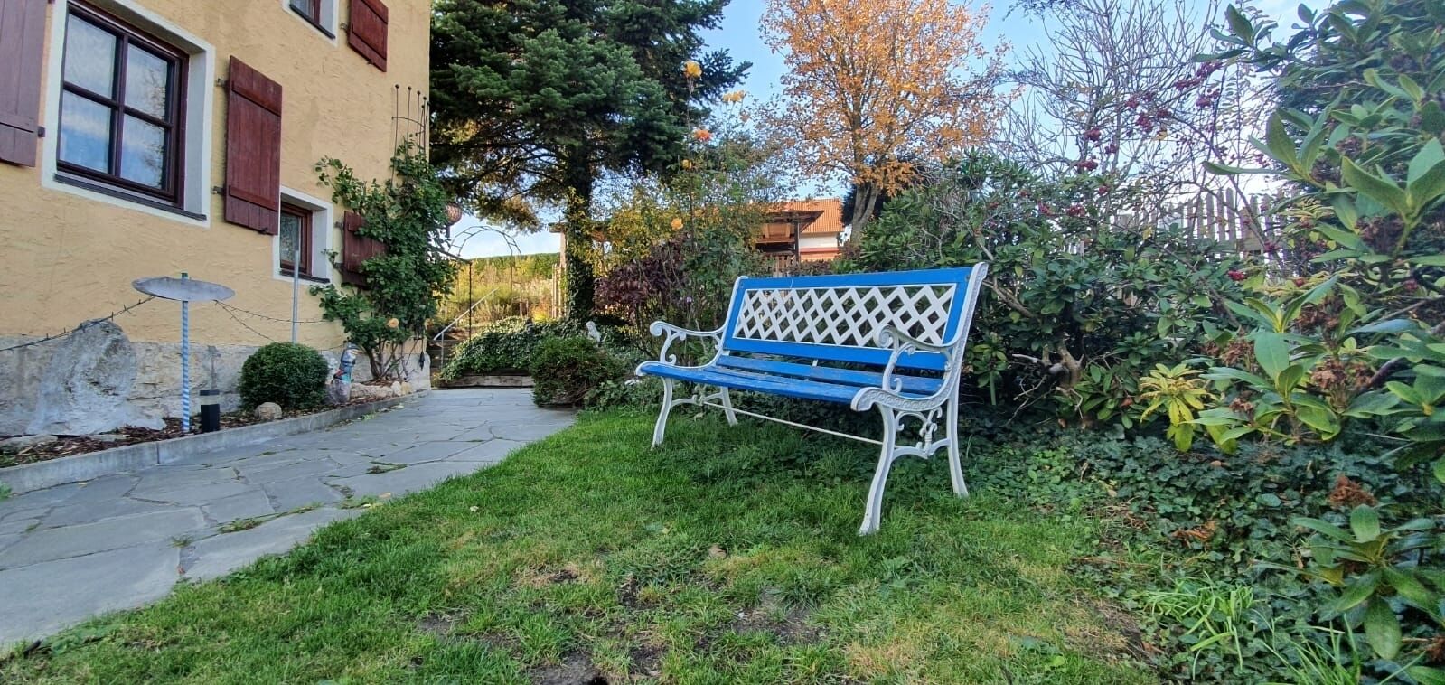 Een bank in een tuin met een pad van stenen dat leidt naar een gebouw met ramen en luiken.