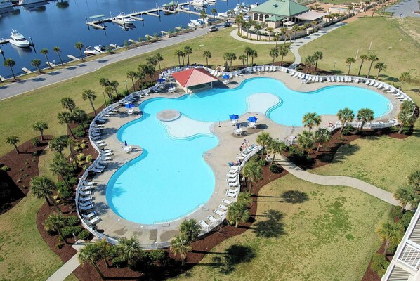 A large, winding pool with lounge chairs and palm trees, surrounded by a marina and a building with a green roof.